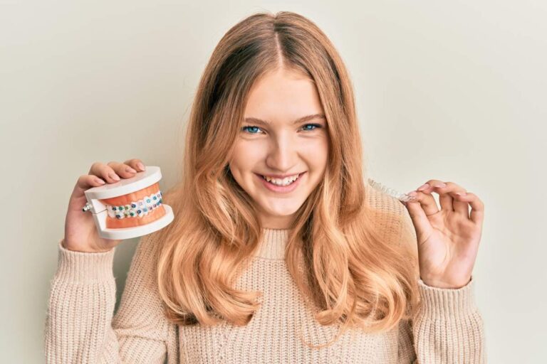 A person holds a clear aligner beside a smiling mouth with neatly arranged teeth.